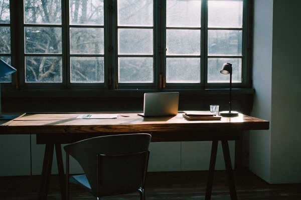 Minimalist workspace with wooden desk, laptop, and desk lamp near large windows.